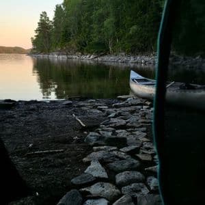 Blick aus dem Zelt auf ein Kanu am felsigen Ufer eines ruhigen Sees vor Waldkulisse bei Sonnenaufgang.