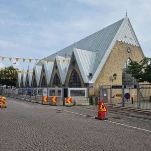 Ein großes Backsteingebäude mit mehreren Spitzgiebeln und einem Metalldach steht an einer Kopfsteinpflasterstraße, die von Bauzäunen gesäumt ist.