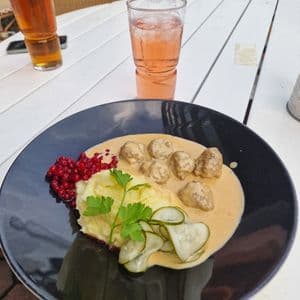 Ein Teller schwedische Köttbullar mit Kartoffelpüree, Preiselbeeren und Gurkenscheiben auf einem weißen Holztisch, dazu Getränke.