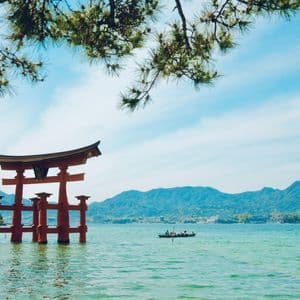 Ein rotes japanisches Torii-Tor steht im Wasser, umrahmt von einem Kiefernast, mit Bergen und einem Boot im Hintergrund.