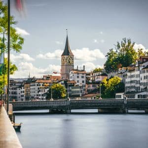 Eine Stadtansicht mit markantem Uhrturm und einer Flussbrücke, von einer baumgesäumten Promenade aus gesehen.