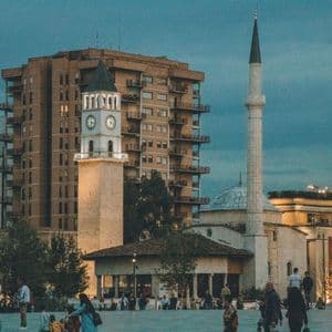 An illuminated clock tower and a mosque's minaret stand in a city square at dusk, with people walking in the foreground.