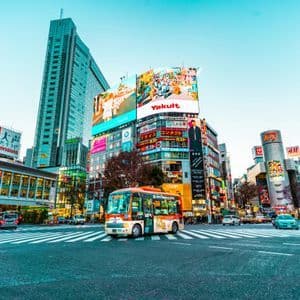 Vue de jour d'une intersection de ville animée avec du trafic traversant devant de grands immeubles recouverts d'enseignes lumineuses et de panneaux publicitaires.