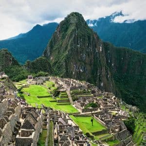 A high-angle view of ancient stone ruins and green terraces nestled in lush mountains, with a prominent peak rising against a cloudy sky.