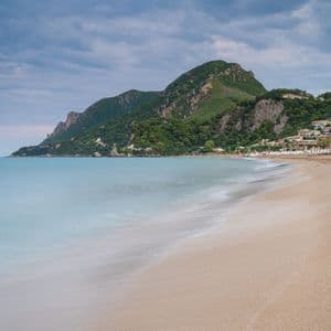 A long-exposure shot of turquoise water washing onto a sandy beach, with a green mountain and coastal buildings in the background.