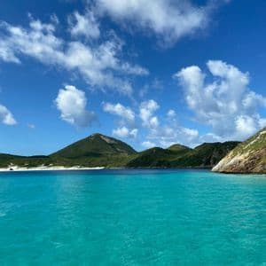 Agua turquesa cristalina en una cala, con colinas verdes y una playa de arena blanca al fondo bajo un cielo azul con nubes.