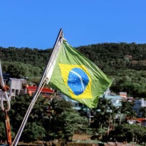 Una bandera de Brasil ondea en un barco, con el telón de fondo de una costa boscosa y un cielo azul.