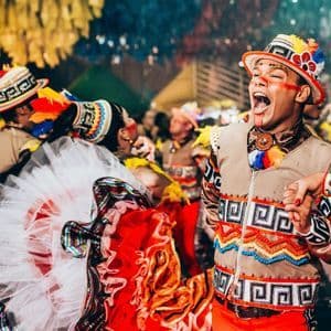 Un hombre con un colorido traje de plumas canta en voz alta mientras baila con una mujer durante un vibrante festival callejero.