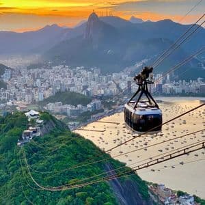 Un teleférico viaja sobre una montaña verde, con vistas a una ciudad, una bahía y montañas al atardecer.
