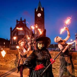 Quattro artisti in costumi elaborati manipolano torce infuocate e oggetti di scena di notte in una piazza cittadina con una torre dell'orologio.