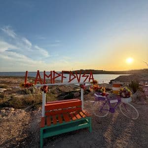 Un grande cartello con lettere arancioni, una panchina colorata e una bicicletta decorativa con fiori su una riva rocciosa al tramonto.