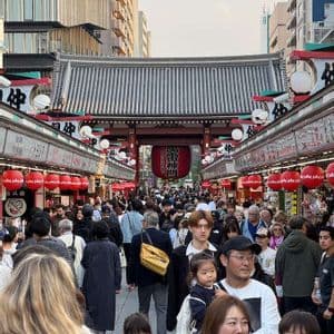 Una fitta folla di persone percorre una tradizionale via di mercato giapponese, fiancheggiata da negozi e lanterne rosse, dirigendosi verso un grande portale.