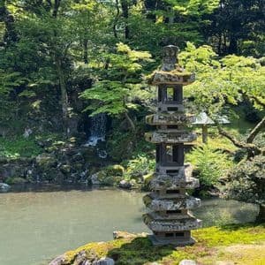 Un'alta lanterna pagoda di pietra si erge su una riva ricoperta di muschio, accanto a uno stagno, in un giardino rigoglioso con una cascata.