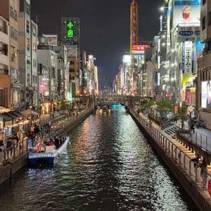 Un canale cittadino di notte, con barche sull'acqua e le sponde fiancheggiate da edifici splendidamente illuminati e insegne al neon.