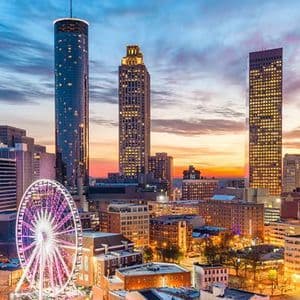 Uno skyline cittadino con alti grattacieli e una grande ruota panoramica illuminata in primo piano contro un cielo colorato al tramonto.