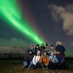 Un viaggio di gruppo WeRoad con abbigliamento invernale, in posa per una foto notturna sotto l'aurora boreale verde e le montagne sullo sfondo.