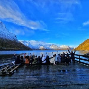 Un gruppo WeRoad è seduto su un molo di legno bagnato con le braccia alzate, di fronte a un fiordo e a montagne innevate.