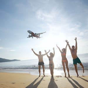 Eine WeRoad-Gruppenreise mit vier Personen, die an einem sonnigen Strand einem tieffliegenden Flugzeug mit erhobenen Armen zuwinken.