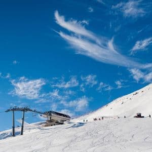 Una seggiovia e un gatto delle nevi su un pendio montano innevato, con sciatori sotto un cielo azzurro brillante e nuvole leggere.