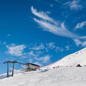 Una stazione di seggiovia su un pendio montano innevato con sciatori e un gatto delle nevi sotto un cielo azzurro brillante con nuvole sottili.