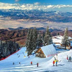 Sciatori su un pendio montano innevato con pini e chalet, che si affaccia su una vasta valle e cime lontane sotto un cielo blu.