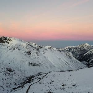 Una cordillera nevada con una carretera serpenteante en el valle bajo un cielo de atardecer rosa y morado.
