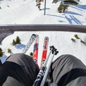 Una perspectiva en primera persona desde un telesilla, con esquís y bastones apoyados en el regazo, con vistas a una ladera de montaña cubierta de nieve.
