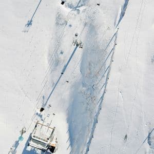 Una vista aérea cenital de un telesilla y su estación sobre un vasto paisaje nevado proyectando largas sombras.