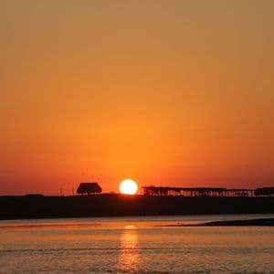 Il sole tramonta all'orizzonte su uno specchio d'acqua, stagliando una costa con diverse piccole capanne di paglia.