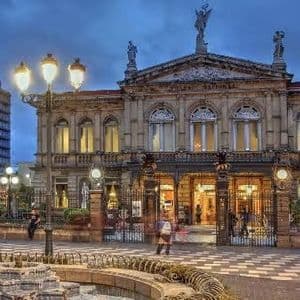 Un sontuoso teatro neoclassico è illuminato su una piazza cittadina al crepuscolo, con una fontana in primo piano.