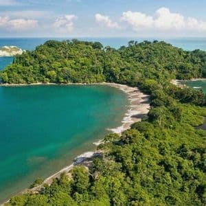 An aerial view of a tropical peninsula covered in dense forest, featuring a curved sandy beach and turquoise ocean water.