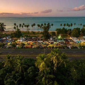 Una veduta aerea di una strada costiera e di una città con negozi illuminati, accanto a una spiaggia con palme al tramonto.