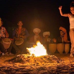 Un grupo de WeRoad se reúne de noche, donde una mujer baila junto a una hoguera mientras unos hombres tocan tambores en la arena.