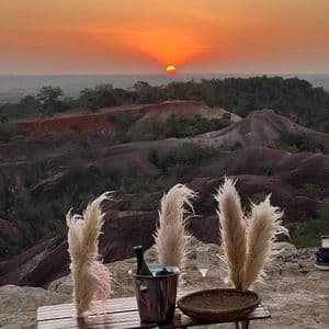 Una bottiglia di vino e bicchieri sono posti su un tavolo con erba della pampas, con vista su un paesaggio collinare al tramonto.