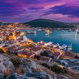 Une ville côtière bien éclairée et son port vus d'un promontoire rocheux au crépuscule sous un ciel violet.