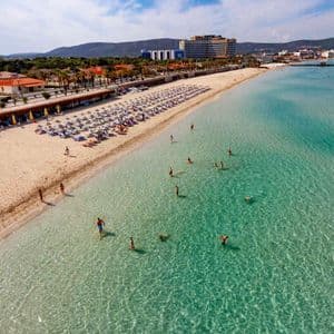 Vue aérienne d'une plage de sable bondée avec des gens nageant dans l'eau turquoise claire et se relaxant sous des rangées de parasols.
