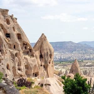 Grandes formations rocheuses avec d'anciennes habitations troglodytiques sculptées, surplombant une vaste vallée avec des collines lointaines.
