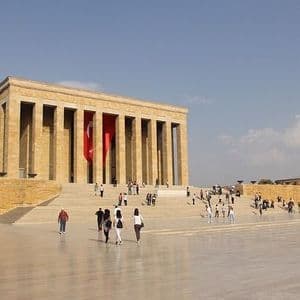 Les visiteurs traversent une grande place en direction d'un imposant bâtiment en pierre avec des colonnes et un grand drapeau turc.