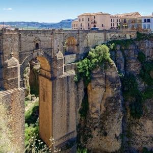 Un grande ponte in pietra con molteplici archi attraversa una gola profonda e rocciosa, con una città arroccata sulle scogliere sotto un cielo azzurro e limpido.