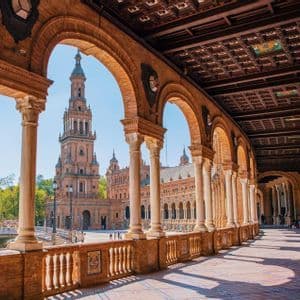 Una vista da una galleria curva con archi e colonne in mattoni che si affaccia su una piazza soleggiata con una torre decorata.