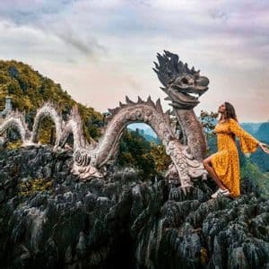 Una donna in abito giallo posa su una cima rocciosa accanto a una grande scultura di drago in pietra.