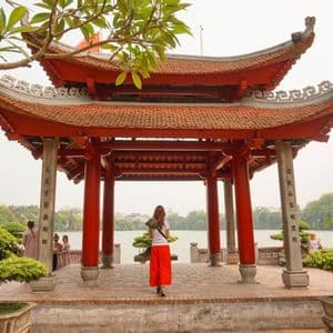 Una donna in pantaloni rossi si trova all'interno di una tradizionale pagoda rossa con tetto a tegole, guardando fuori su un lago con alberi e bonsai.