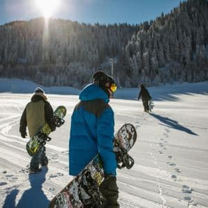 Un viaggio di gruppo WeRoad con tre persone che, portando gli snowboard, camminano su un pendio montano soleggiato e innevato.