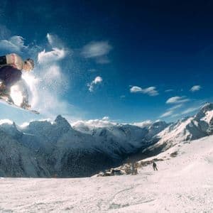 Uno snowboarder salta in aria sopra una catena montuosa innevata sotto un cielo azzurro chiaro.