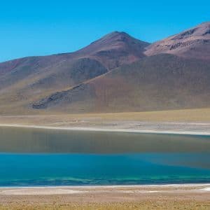 Un lago calmo e turchese si trova alla base di montagne aride e rossastre sotto un cielo azzurro e limpido.