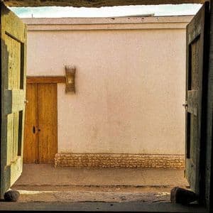 Vista da un interno buio attraverso porte di legno aperte su un cortile soleggiato con un muro imbiancato e un'altra porta.