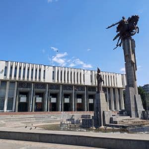 Una estatua ecuestre monumental y una fuente en una plaza frente a un gran edificio blanco bajo un cielo azul claro.
