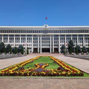 A large government building with a flag on top, viewed from across a plaza with fountains and colorful flowerbeds.