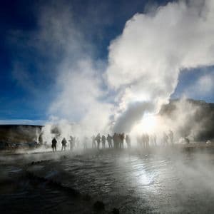 Sagome di un gruppo WeRoad tra geyser in eruzione, con vapore che si innalza contro un sole splendente e un cielo blu.