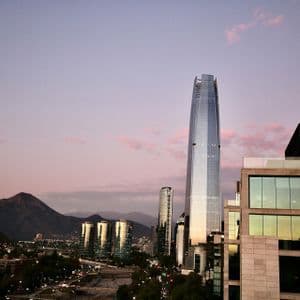Uno skyline cittadino moderno con diversi grattacieli che si stagliano su una catena montuosa sotto un cielo rosa e viola al tramonto.